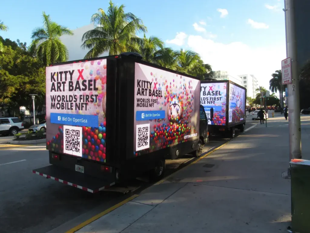 Mobile billboard trucks advertising "Kitty X Art Basel" and "World's First Mobile NFT" with colorful graphics and QR codes, set against a palm tree-lined street, illustrating innovative guerrilla marketing strategies.