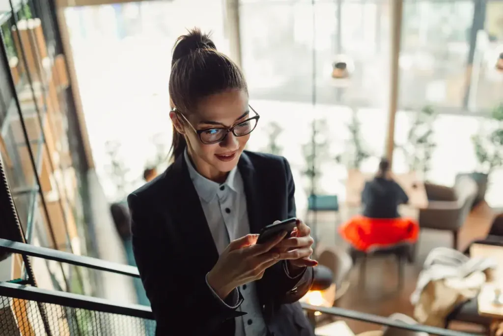 Professional woman in glasses using smartphone in modern office setting, reflecting themes of marketing strategy and digital engagement.