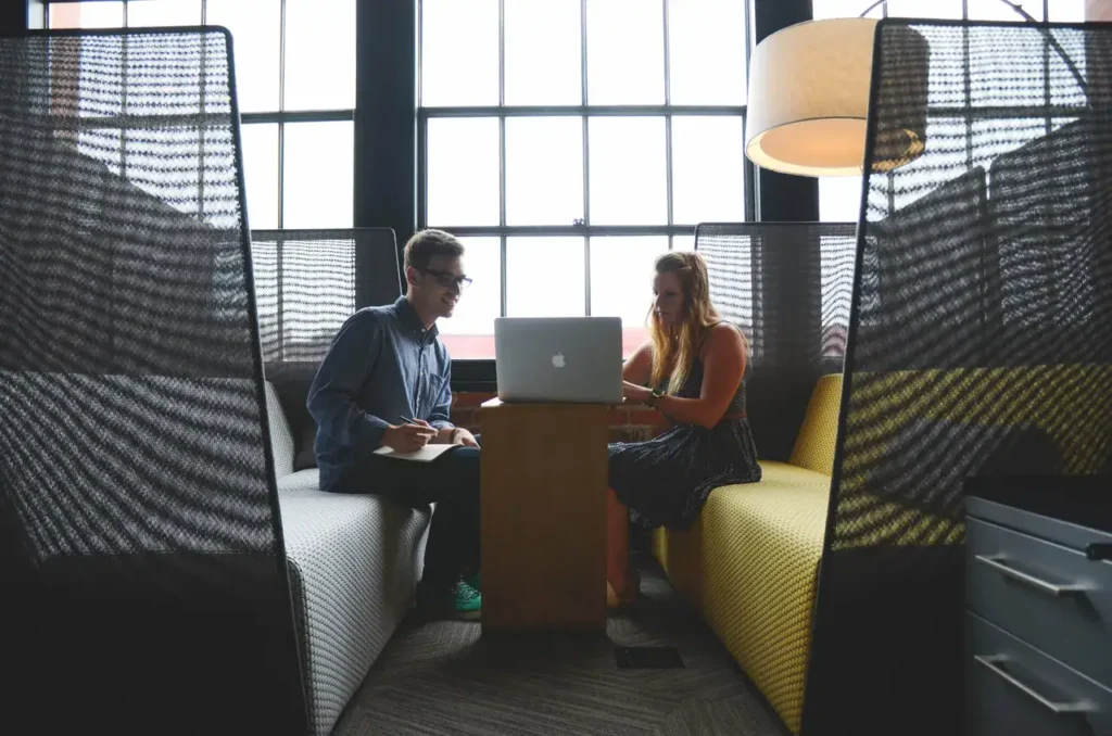 Two professionals collaborating in a modern workspace, discussing ideas over a laptop at a shared table, with cozy seating and large windows in the background.