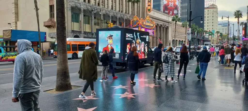 Crowd of pedestrians on Hollywood Boulevard near LED mobile billboard advertising, showcasing dynamic content amid urban scenery and palm trees.