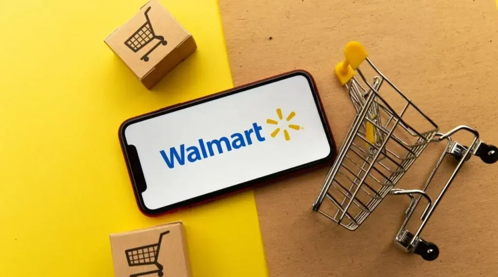 Walmart logo on smartphone next to miniature shopping cart and boxes, representing in-store advertising and e-commerce strategies.