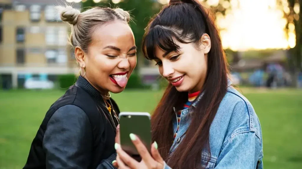 Two young women smiling and interacting while looking at a smartphone in a park, representing social engagement and modern communication, relevant to event staffing and marketing strategies.