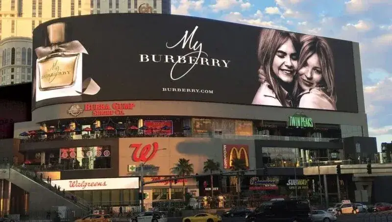 Large billboard advertisement for Burberry perfume featuring two smiling women, located on a busy street with storefronts like Walgreens and McDonald's visible below.