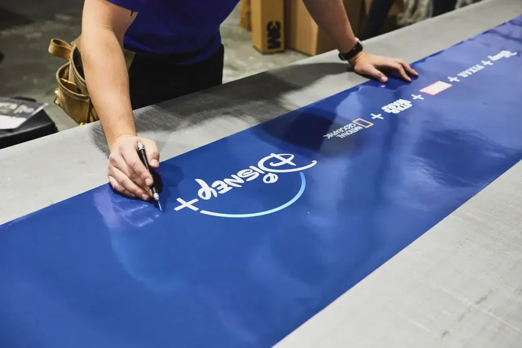 Person writing on a blue banner featuring the Disney+ logo, showcasing creative branding for guerrilla marketing.