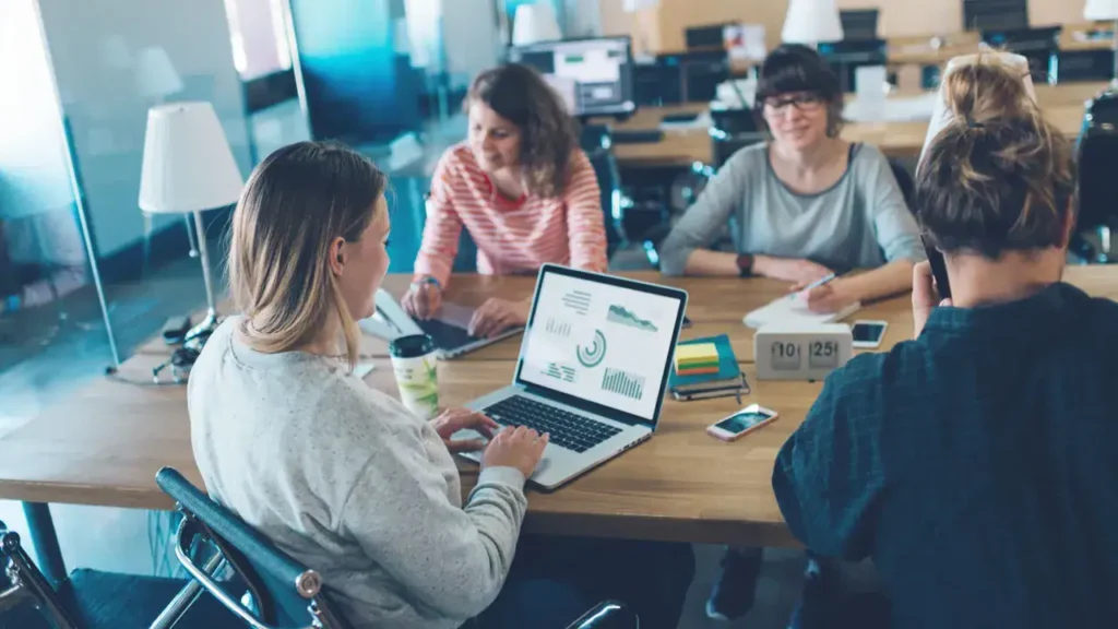 Group of diverse professionals collaborating at a table with a laptop displaying marketing data and graphs, emphasizing teamwork and brand strategy in a modern office setting.