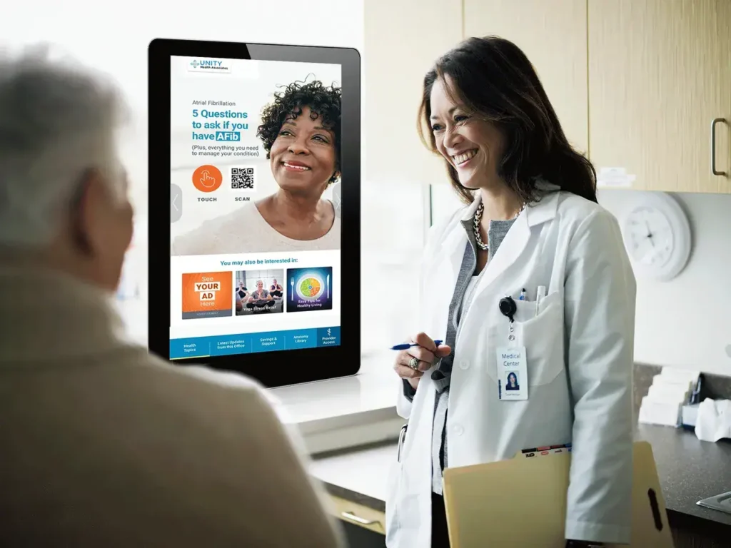 Doctor discussing health information with patient, digital display showing health-related advertisement and QR code, medical office setting.