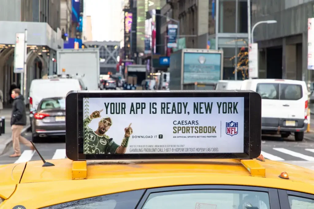 Taxi advertising featuring "YOUR APP IS READY, NEW YORK" for Caesars Sportsbook, highlighting mobile app promotion amidst a busy urban backdrop.