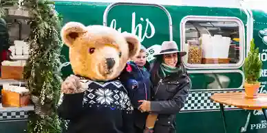Person in a bear mascot costume with a child and woman, standing in front of a green food truck, promoting a festive atmosphere at an event.