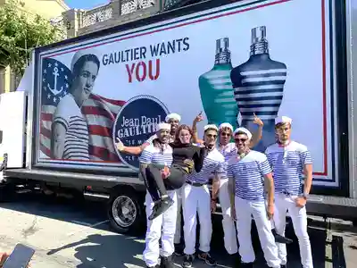 Brand ambassadors posing in front of a promotional truck for Jean Paul Gaultier, featuring bold branding and an American flag backdrop, showcasing vibrant marketing strategies in an urban setting.