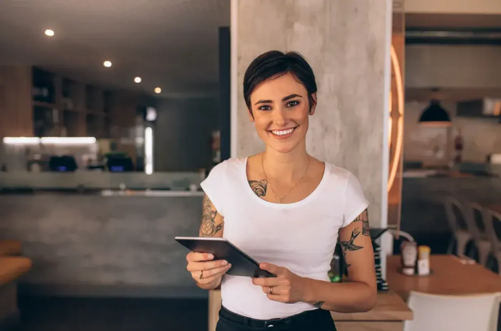 Smiling woman with tattoos holding a tablet in a modern caf&eacute; setting, representing event staffing and brand ambassador services by American Guerrilla Marketing.