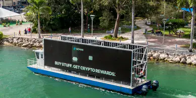 Billboard advertising on a boat promoting cryptocurrency rewards, featuring the text "BUY STUFF, GET CRYPTO REWARDS," set against a waterfront park in Las Vegas.