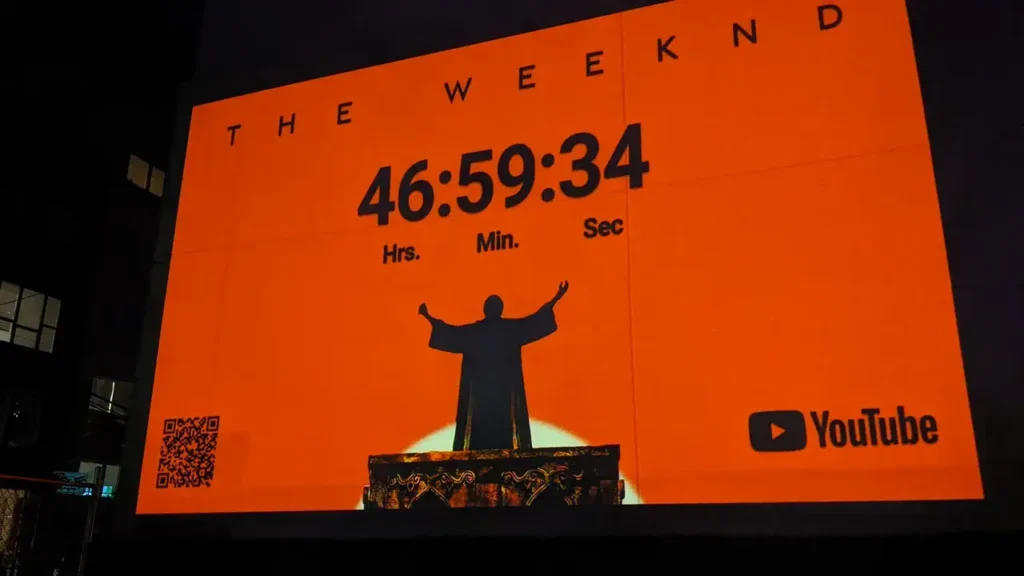 Countdown timer for The Weeknd's event displayed on a large orange backdrop, featuring a silhouetted figure with outstretched arms, alongside YouTube logo and QR code.