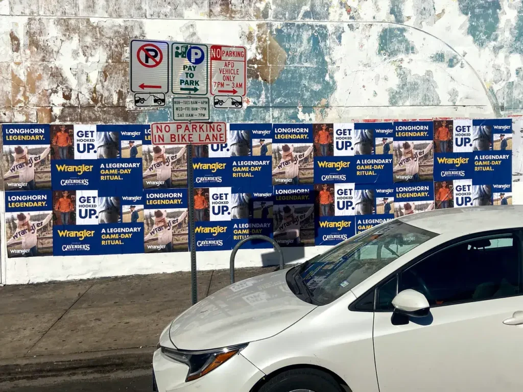 White car parked near advertising posters for Wrangler and Longhorn, featuring phrases "GET UP" and "A LEGENDARY GAMEDAY RITUAL," in a vibrant urban setting.