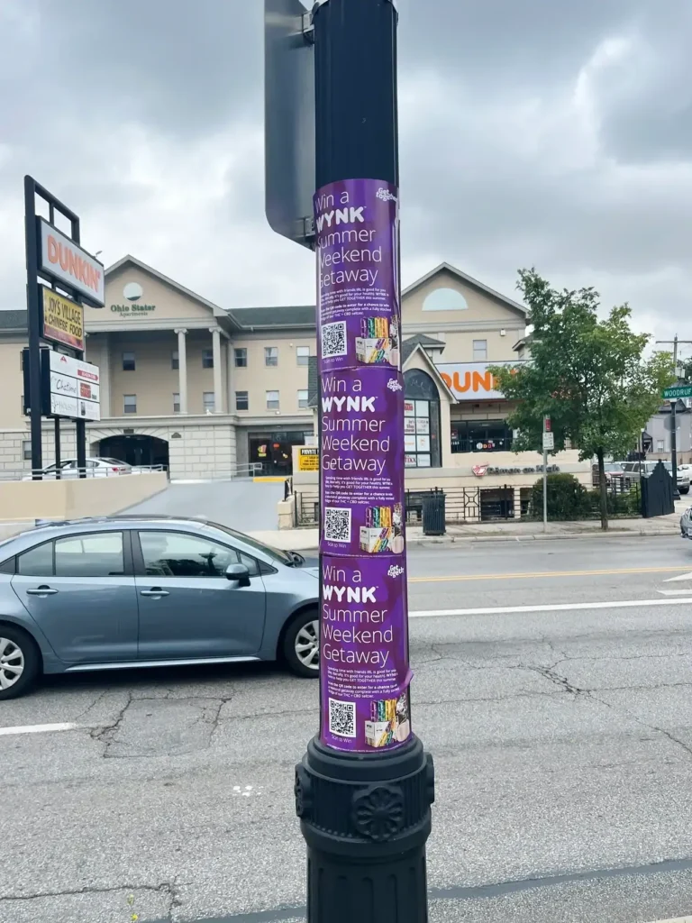 Purple promotional posters on a lamppost advertising a "WYNK Summer Weekend Getaway," featuring QR codes and colorful graphics, with a Dunkin' store in the background.