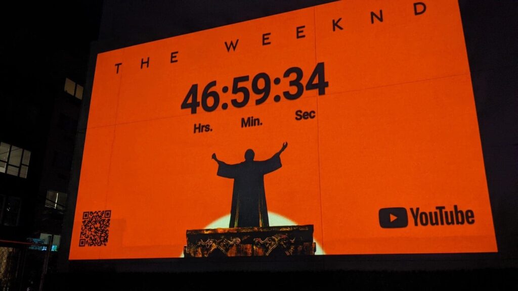 Projection advertising for The Weeknd, featuring a countdown timer and silhouette figure against an orange background, with YouTube logo and QR code.