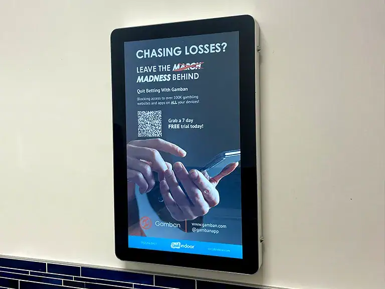 Digital advertisement for Gamban promoting responsible gambling, featuring hands holding a smartphone with a QR code, displayed in a restroom setting, emphasizing the message to quit betting and providing a free trial offer.