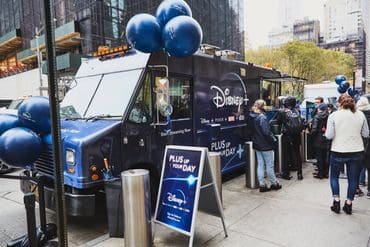 Disney-branded food truck with blue balloons, people lined up, and promotional signage for Plus Day, illustrating innovative bus advertising strategies.