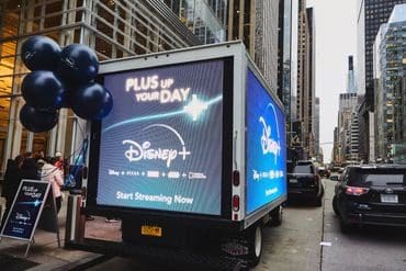 Disney+ promotional truck with digital billboard displaying "Plus Up Your Day" and "Start Streaming Now," surrounded by balloons, in urban setting.