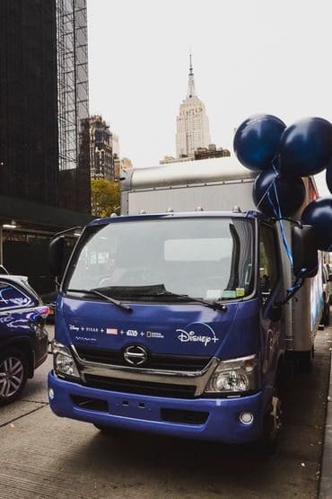 Disney+ branded LED billboard truck with blue balloons parked in an urban setting, featuring the Empire State Building in the background, representing innovative outdoor advertising solutions.