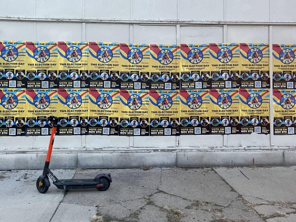 Wall covered in colorful election posters promoting voting with a scooter parked in front, emphasizing grassroots engagement and guerrilla marketing strategies.