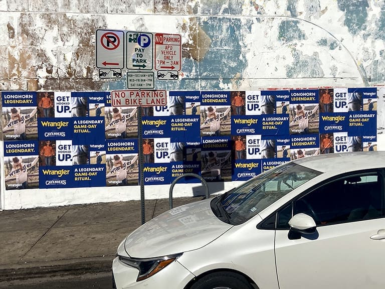 White car parked near promotional banners for Wrangler and Longhorn, featuring "GET UP!" messaging, with parking signs indicating regulations.