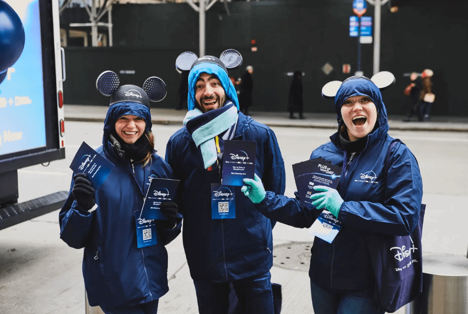 Three enthusiastic brand ambassadors wearing Disney-themed attire and mouse ears, holding promotional materials, engaging with passersby in a city street setting.