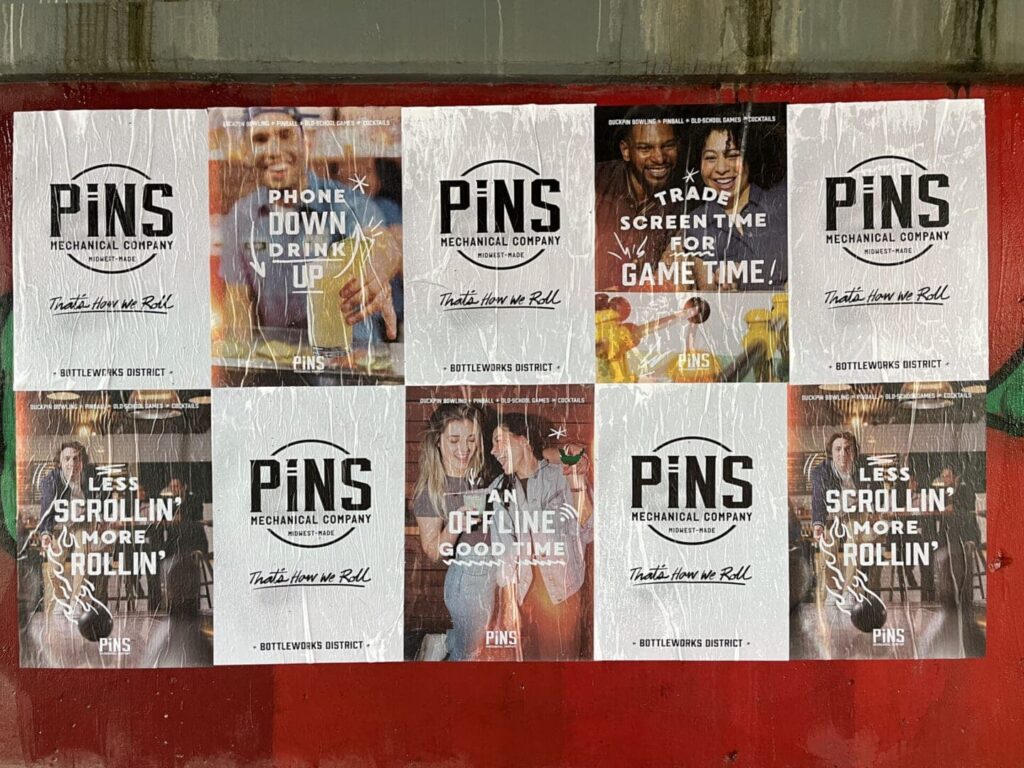 Promotional posters for PiNS Mechanical Company featuring slogans encouraging social interaction and fun activities, with messages like "Phone Down, Drink Up" and "Trade Screen Time for Game Time," displayed in the Bottlenworks District.