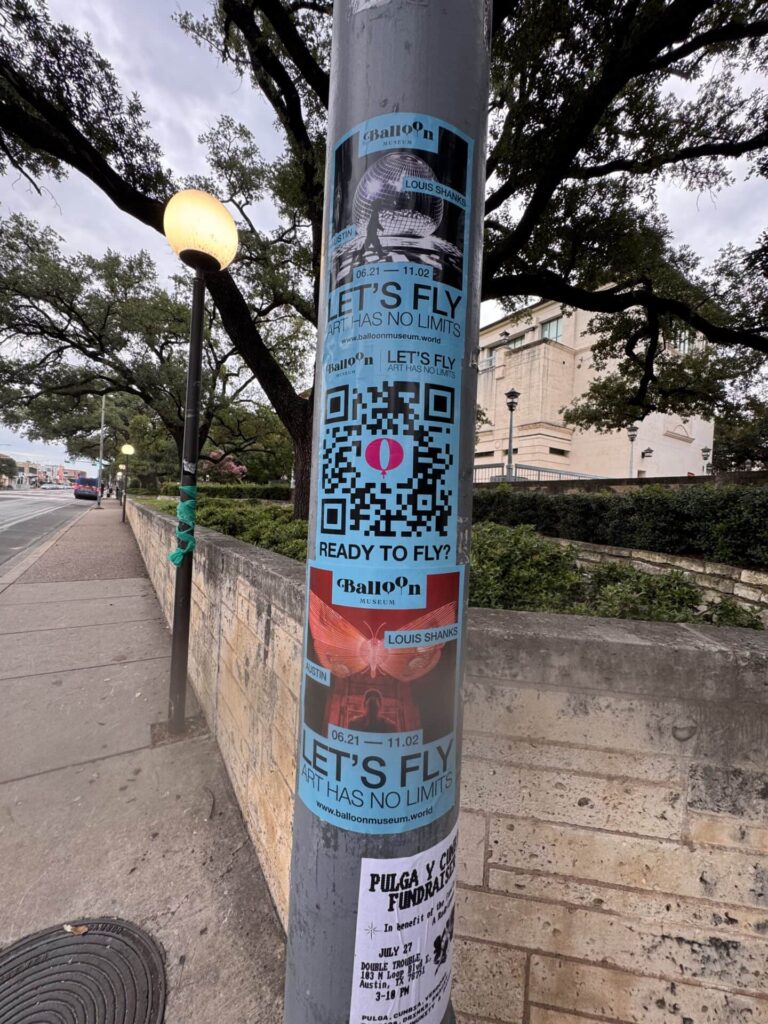 Billboard advertising on a street pole featuring colorful flyers for "Balloon" with QR code, promoting an event with the slogan "LET'S FLY," emphasizing limitless experiences, relevant to guerrilla marketing strategies.