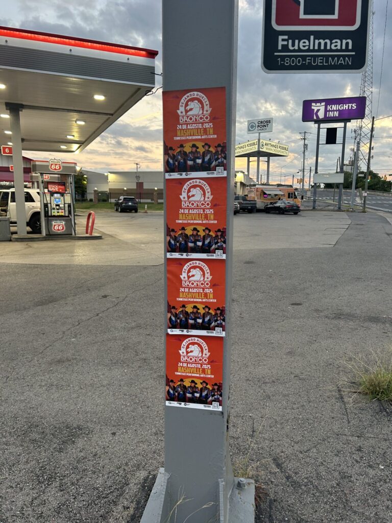 Posters promoting an event featuring a group of people, displayed on a pole at a gas station, with a sunset backdrop and nearby signage indicating fuel services.