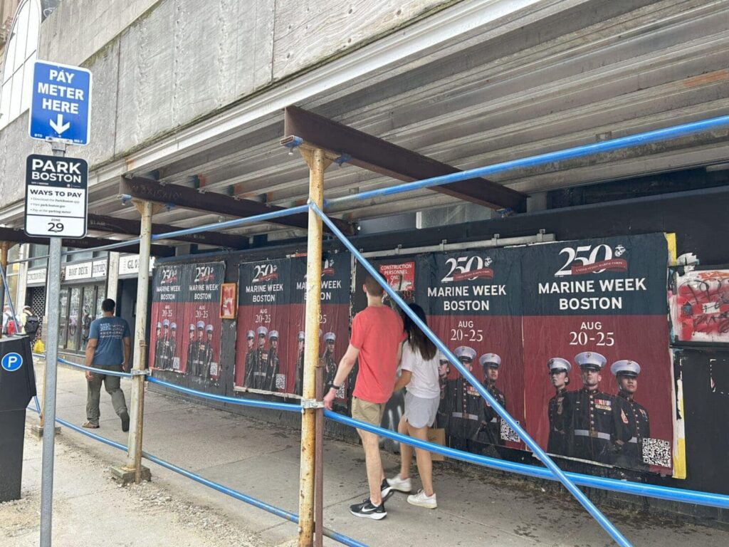 Street scene featuring pedestrians walking past construction barriers displaying promotional posters for Marine Week Boston, featuring dates August 20-25 and images of Marines, with parking meter signage.