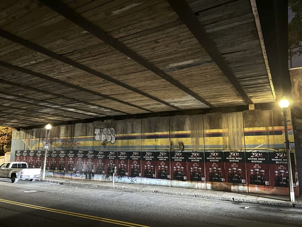 Urban marketing wall featuring promotional posters for college events and activities, illuminated by streetlights, under a wooden overpass.