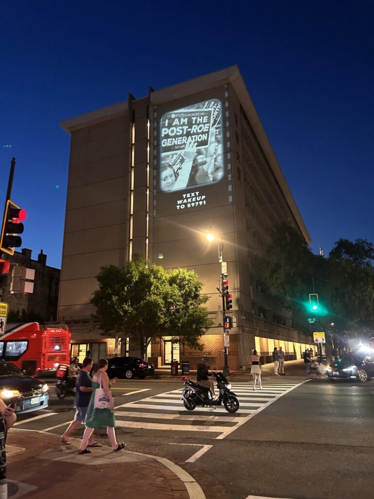 Projection on building reading "I AM THE POST-ROE GENERATION" with call to action, urban street scene, pedestrians crossing, nighttime setting.