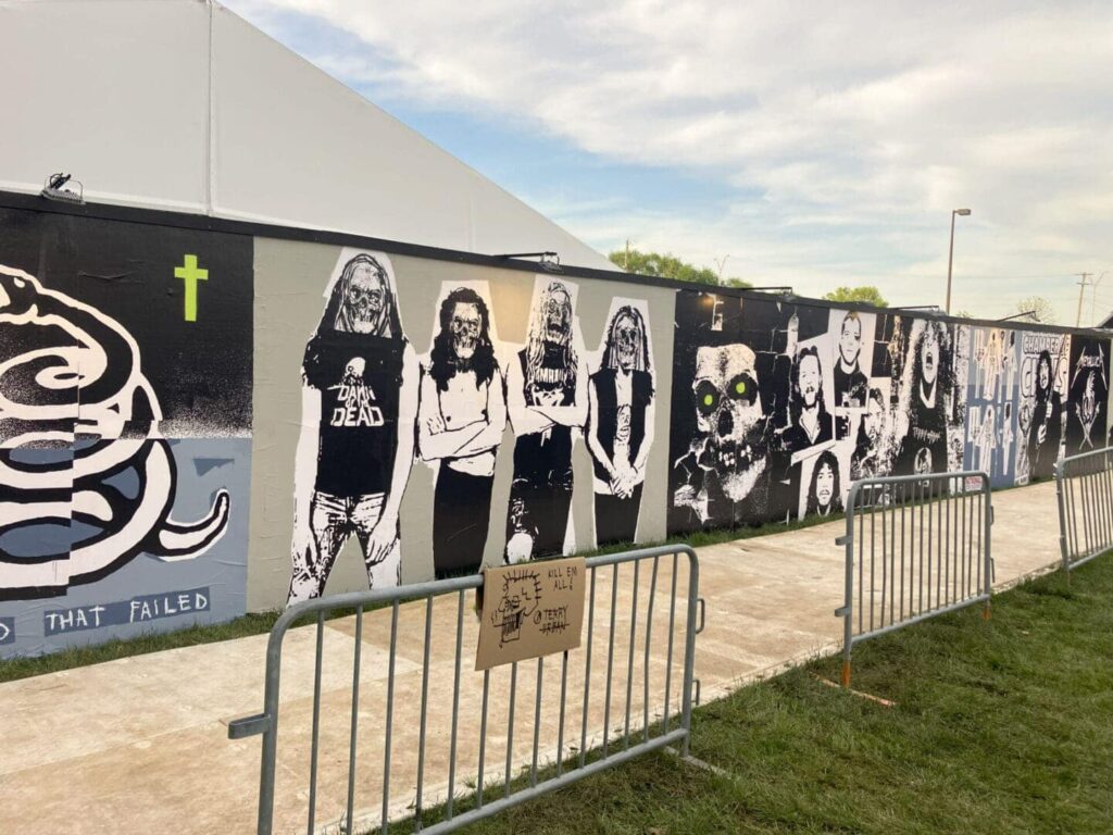 Mural featuring rock band imagery and graffiti art, showcasing various band members and iconic symbols, with barriers in front, set in an outdoor festival environment.
