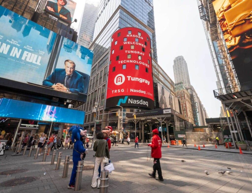 Billboard at Nasdaq Tower in Times Square displaying "NASDAQ WELCOMES TUNGRAY" and colorful advertisements, with people in the foreground engaging in the vibrant urban setting.