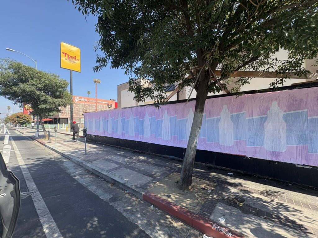 Colorful mural on a wall featuring abstract figures, with a tree and sidewalk in foreground, set in an urban environment near a fast-food restaurant.