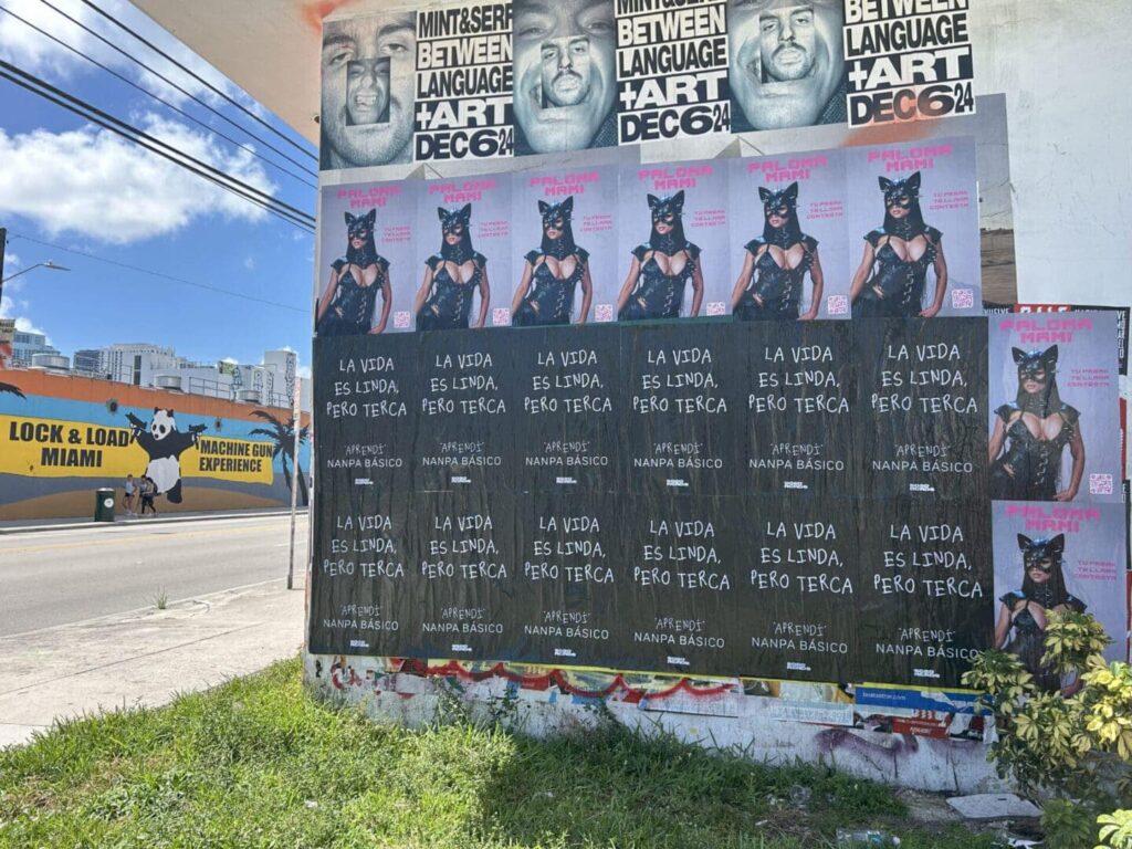 Posters featuring a woman in a cat costume with text "LA VIDA ES LINDA, PERO TERCA" promoting a performance by Nampa Basico, surrounded by urban graffiti and advertisements in Miami.