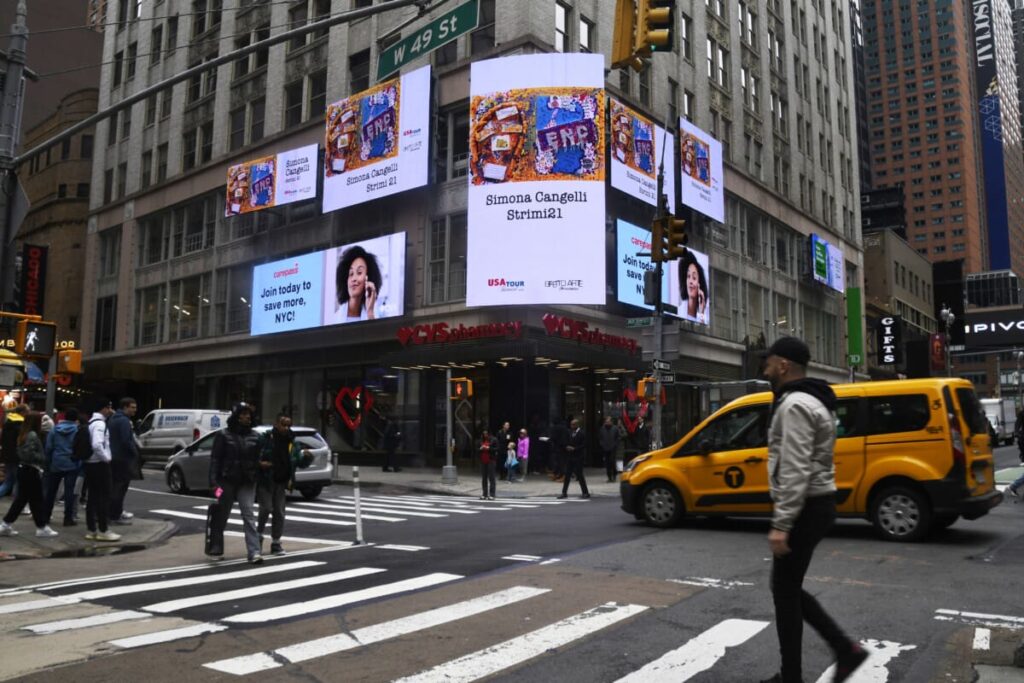 Times Square digital billboards displaying advertisements, including a promotional message and an image of a woman, with pedestrians and a yellow taxi in the foreground.