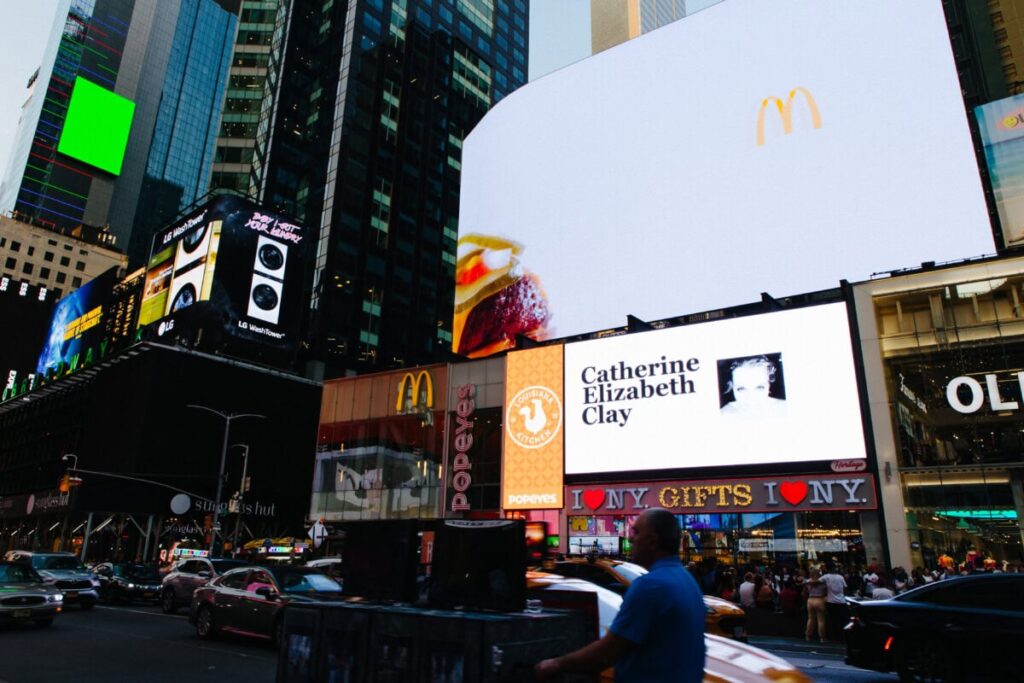 Billboard advertising in Times Square featuring McDonald's logo and a personal message for Catherine Elizabeth Clay, surrounded by bustling city activity and other advertisements.