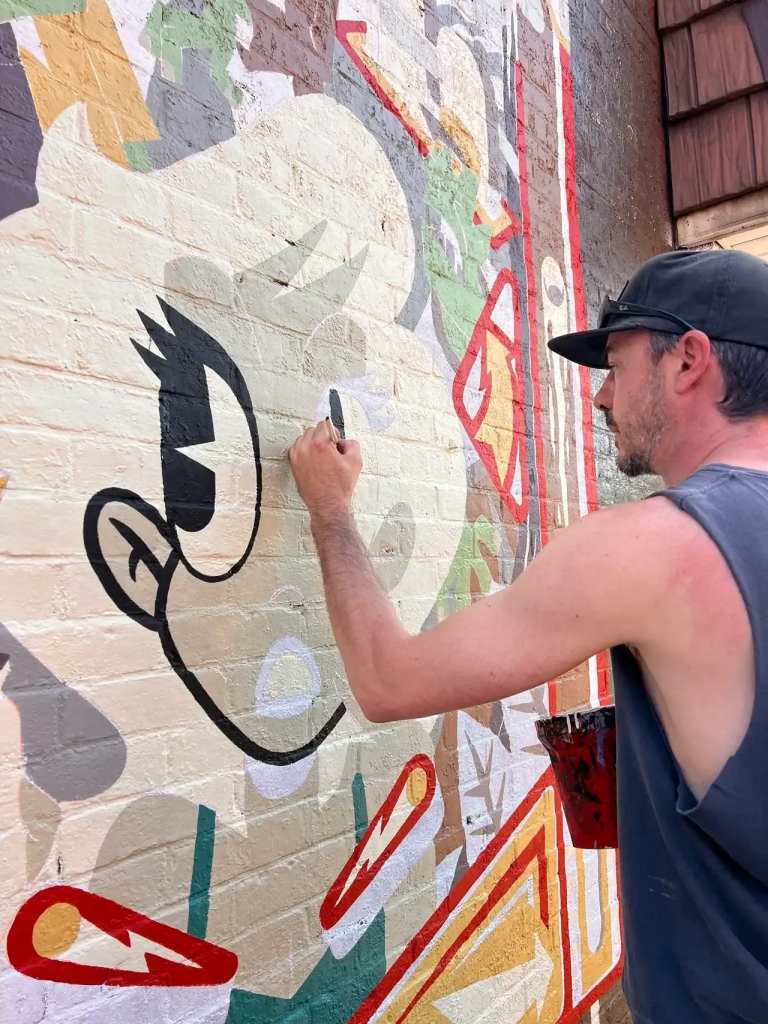 Artist painting a colorful wall mural featuring a stylized character, showcasing outdoor mural advertising techniques relevant to American Guerrilla Marketing services.