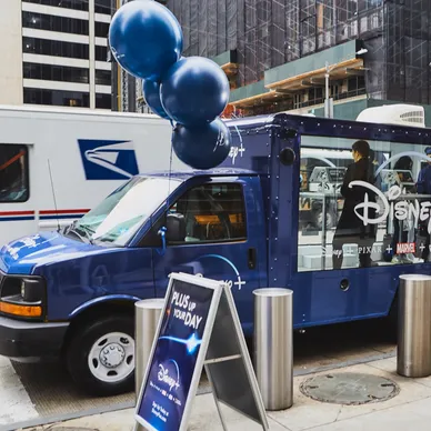 Mobile billboard truck promoting Disney+ with blue balloons, parked in an urban setting, featuring branding and a promotional sign.