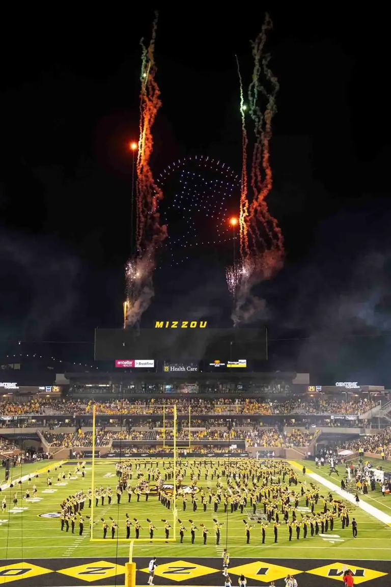 Fireworks display above a stadium with "MIZZOU" sign, featuring a marching band formation on the field, enhancing the vibrant atmosphere of a drone light show event.