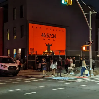 Large illuminated billboard displaying a countdown timer for The Weeknd, with pedestrians walking by in a city street at night.