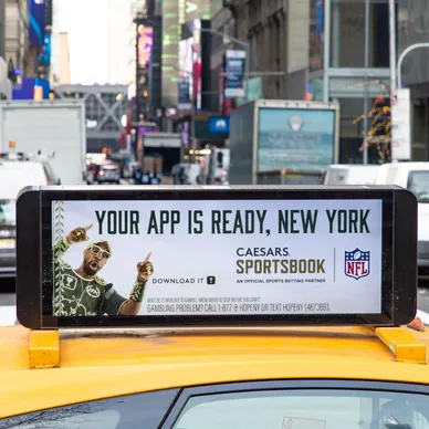 Taxi top advertisement featuring a promotional message for Caesars Sportsbook, highlighting "YOUR APP IS READY, NEW YORK" with an image of a smiling man, set against an urban backdrop in a busy street environment.
