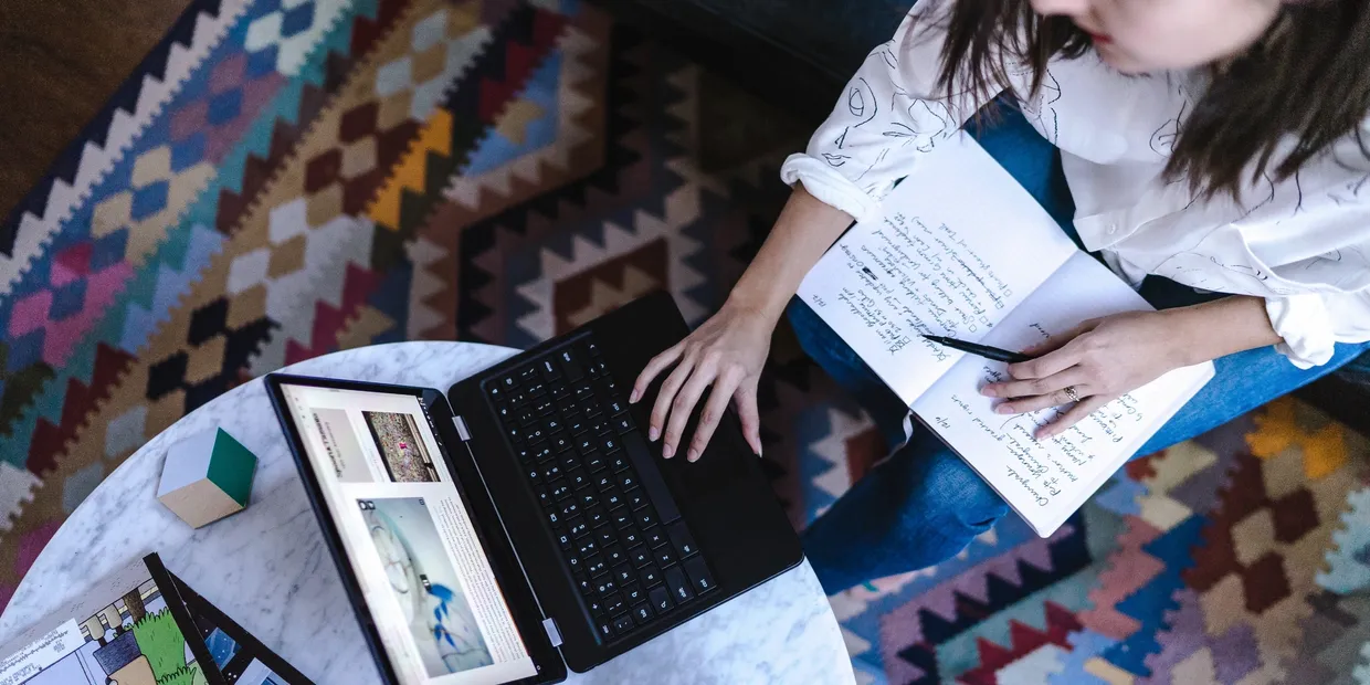 Person working on a laptop with notes on a marble table, surrounded by a colorful patterned rug, illustrating remote work and productivity.
