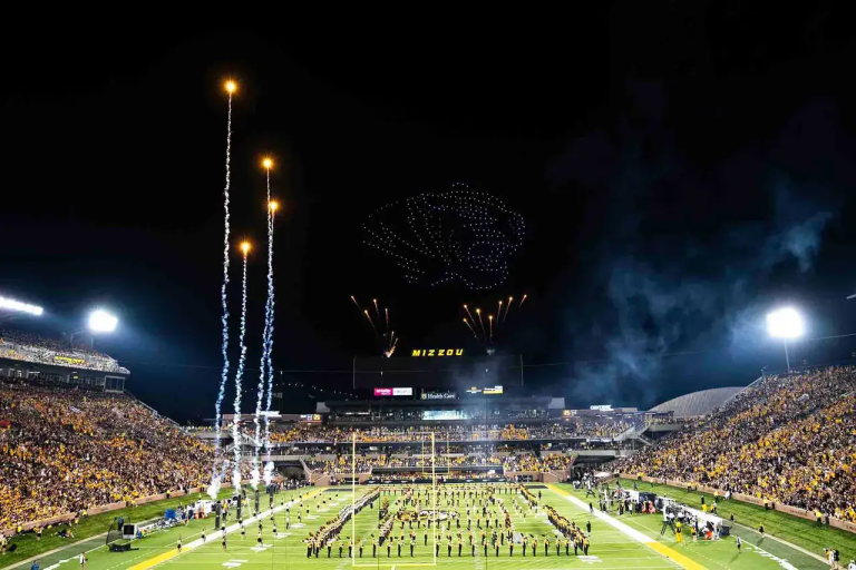 Drone light show featuring aerial displays and fireworks over a packed stadium during a night event, with a formation of drones creating a lion shape and the word "MIZZOU" illuminated in the background.