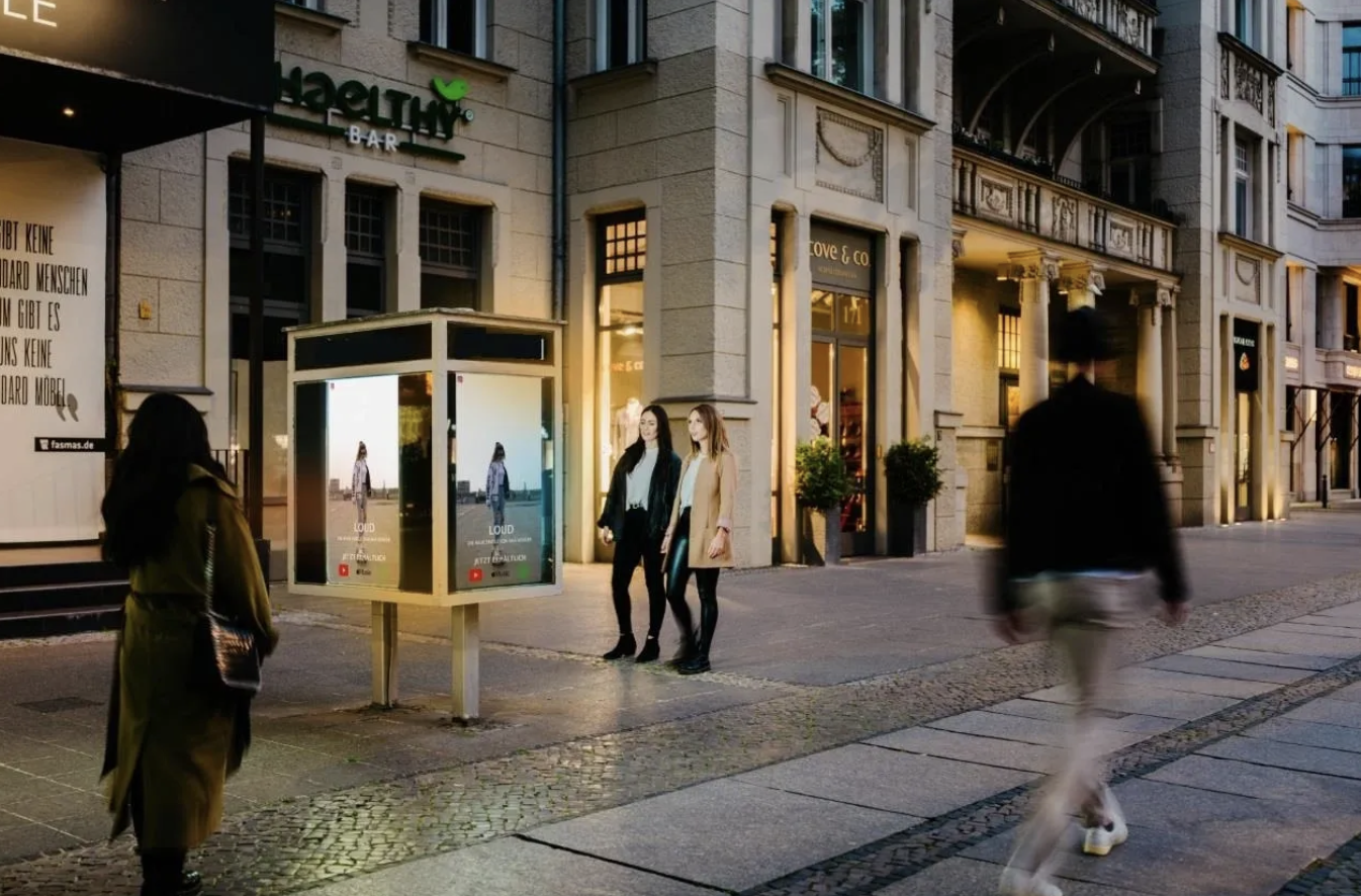 Two women standing by a digital billboard in an urban setting, showcasing outdoor advertising, with pedestrians walking past and shops in the background, highlighting international billboard advertising strategies.