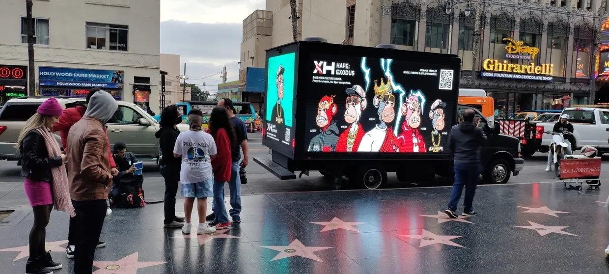 Billboard advertising truck displaying vibrant animated characters in a busy urban setting, with pedestrians engaged nearby on the Hollywood Walk of Fame.