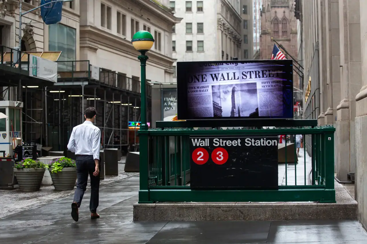 Man walking towards Wall Street subway station with digital advertisement displaying "One Wall Street" in a bustling urban setting, emphasizing subway advertising's potential for visibility and engagement.