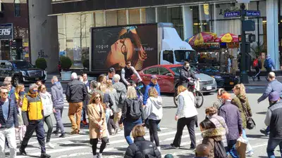 Mobile billboard truck displaying a fashion advertisement in a busy urban crosswalk with pedestrians walking and vehicles in the background.