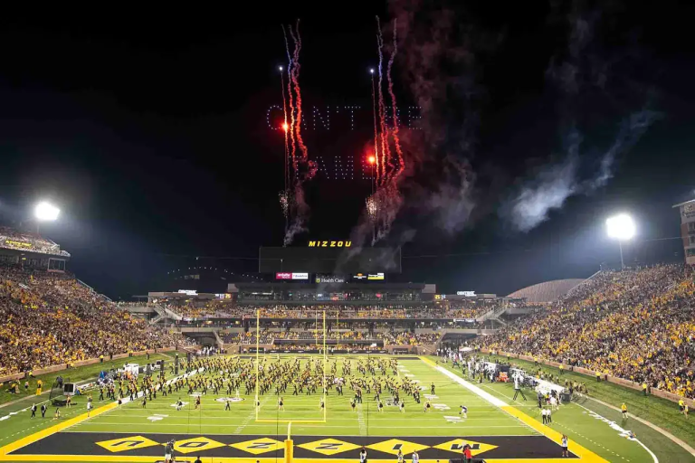 Fireworks display above Mizzou stadium during a drone light show, with a vibrant crowd on the field celebrating an event.